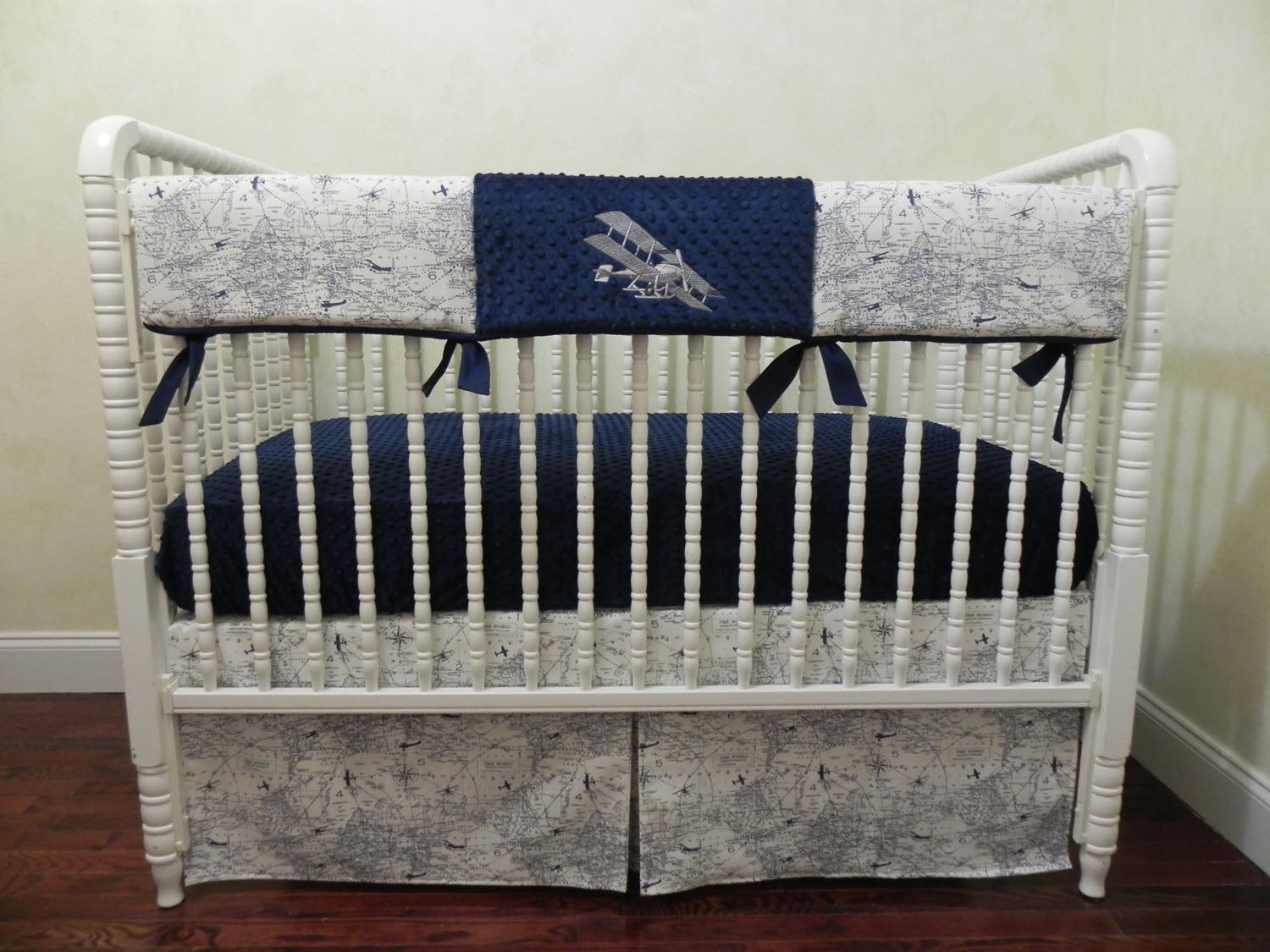 A white baby crib with airplane-themed bedding, including a crib sheet with air traffic map print, a rail cover with a bi-plane design, and an accent pillow.