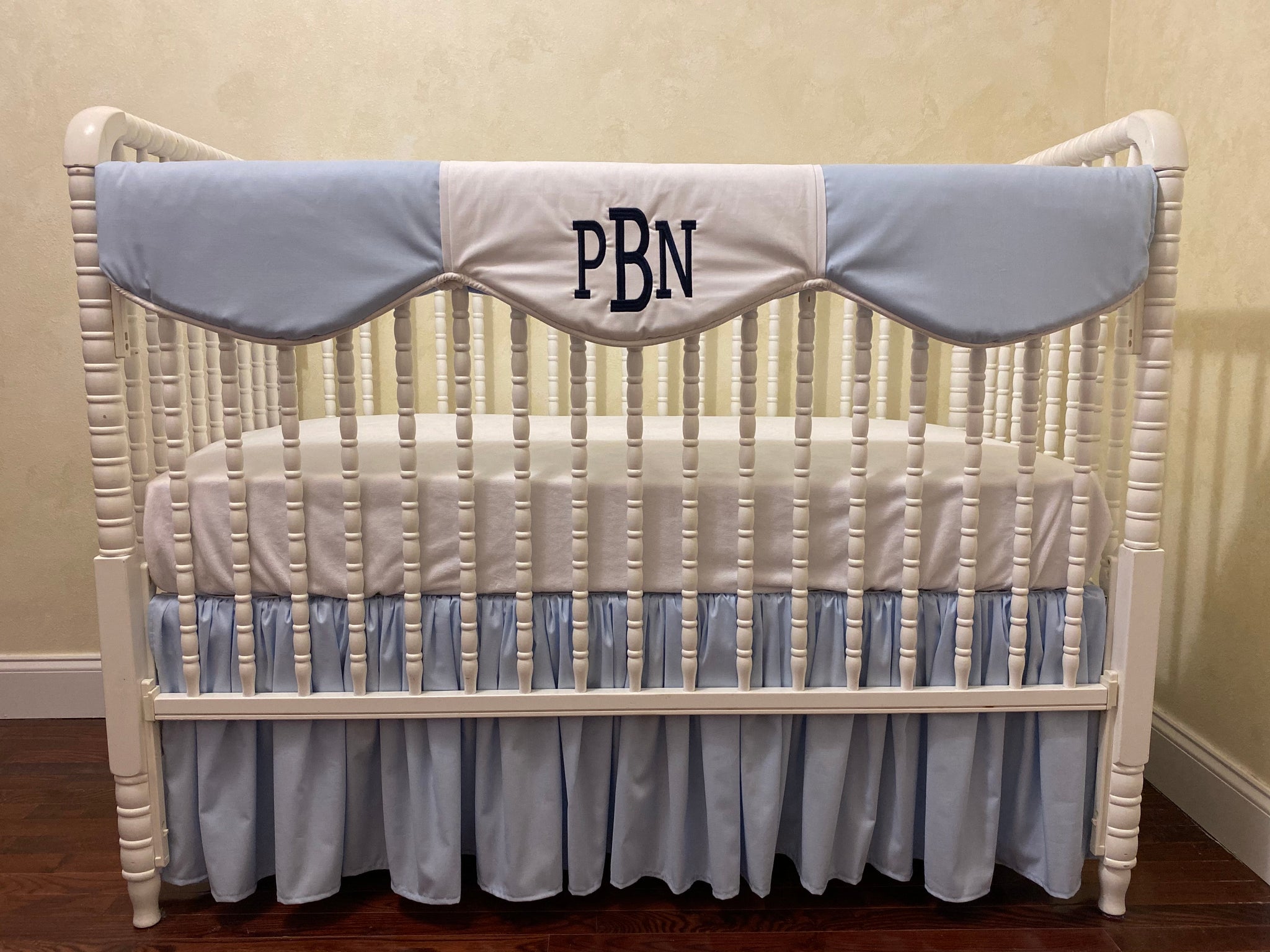 A crib with light blue and white bedding, including a scalloped rail guard and a navy monogram on the crib rail cover.