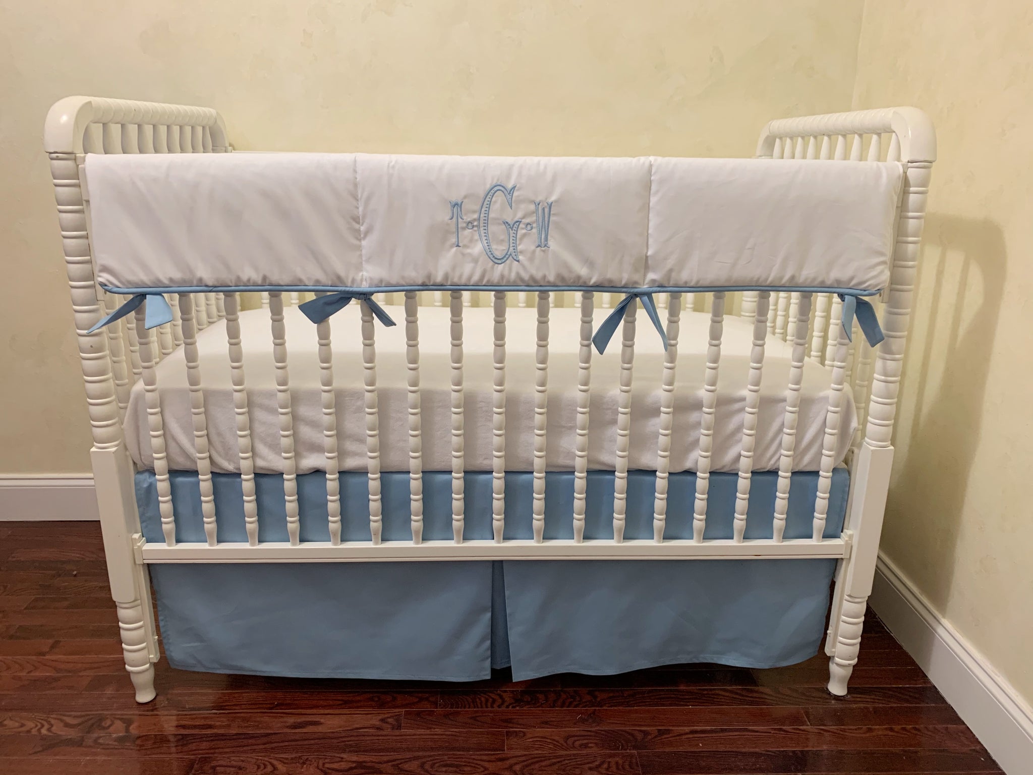 A crib with white rail cover and light blue accents, featuring a personalized monogram on the cover, placed in a room with a wooden floor and a light blue wall.