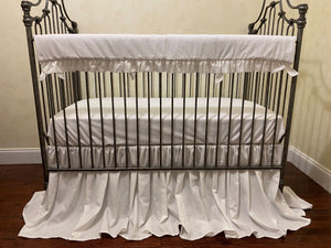 Iron crib with white bedding and skirt on a wooden floor.