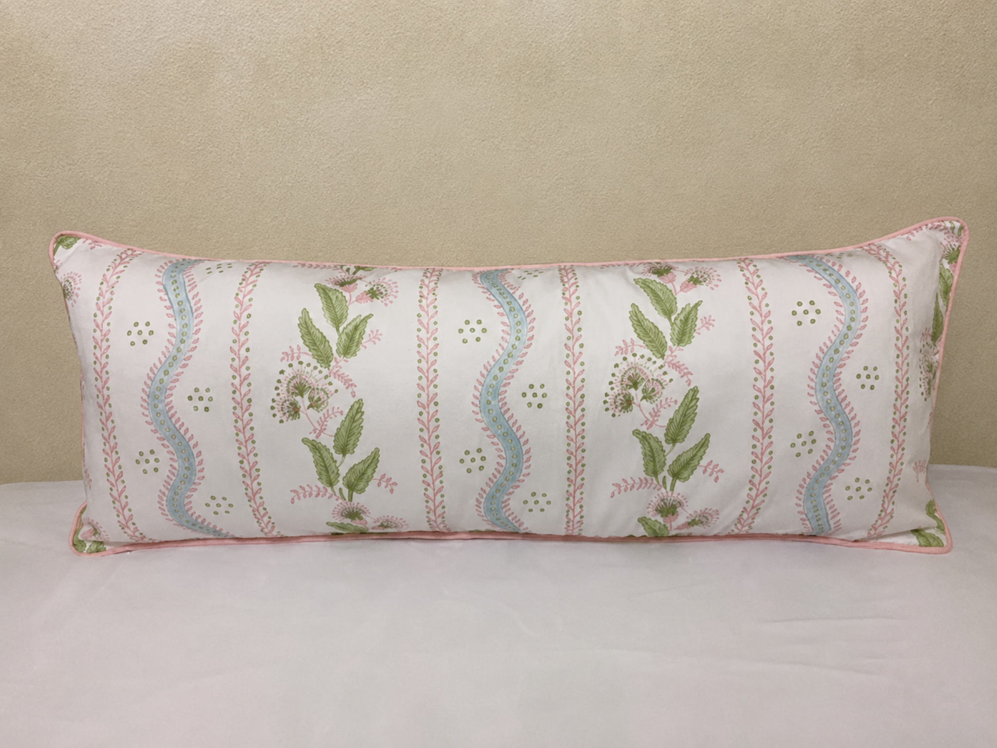 A rectangular lumbar pillow with a white background, featuring vertical stripes of pink floral embroidery, green leaves, and light blue wavy patterns, finished with a pink piped edge.